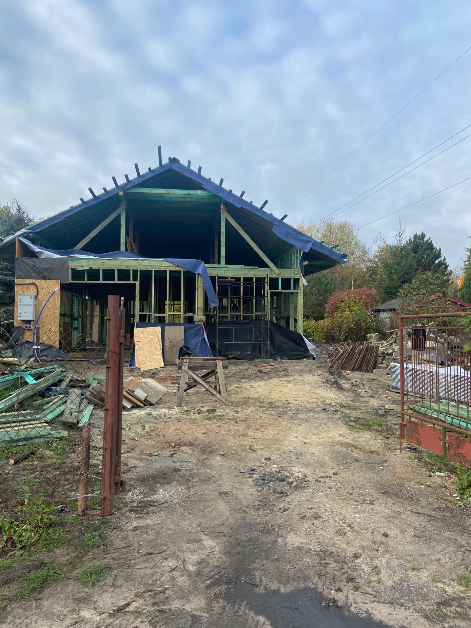 Konstrukcja drewnianego domu w trakcie budowy, zabezpieczona folią, widoczne elementy szkieletu i instalacje elektryczne, teren budowy z materiałami i narzędziami.