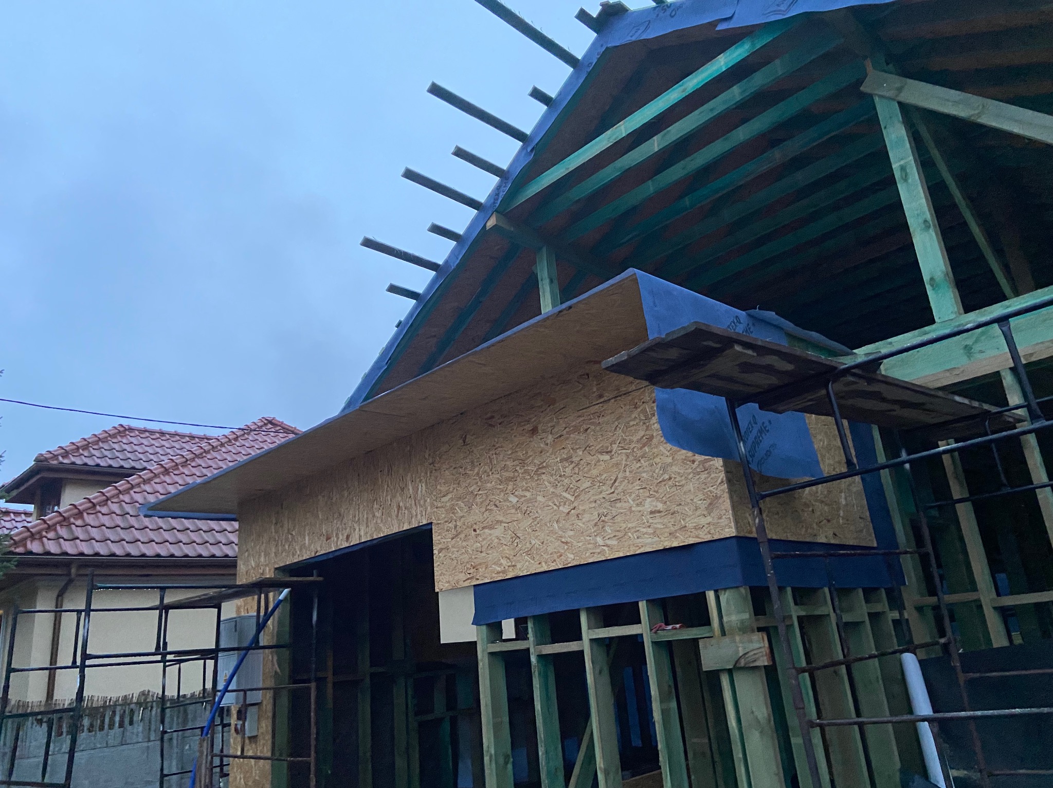 Wznoszenie domu w technologii szkieletowej: widok na konstrukcję z zielonych belek, płyt OSB i niebieskiej membrany dachowej, z rusztowaniem w pierwszym planie i fragmentem sąsiedniego domu...