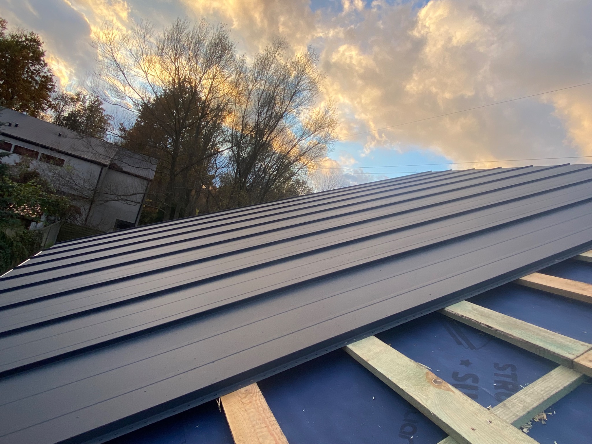 Montaż blachy na dachu z widocznymi łatami i membraną dachową, na tle zachmurzonego nieba i drzew.