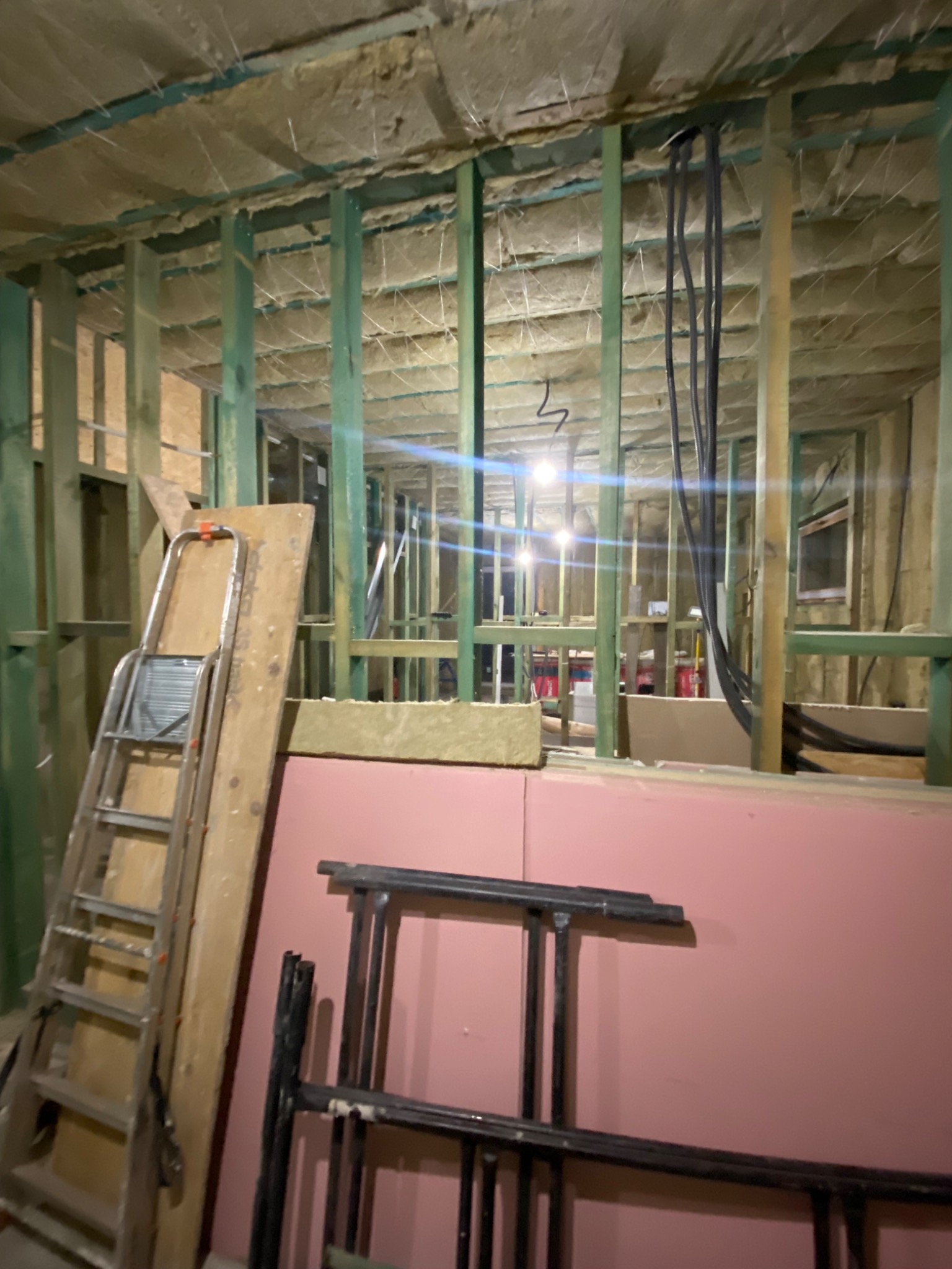 Wnętrze domu szkieletowego w trakcie budowy z widoczną izolacją stropu, ścianami z drewnianej ramy, instalacją elektryczną, opartą drabiną i rusztowaniem.