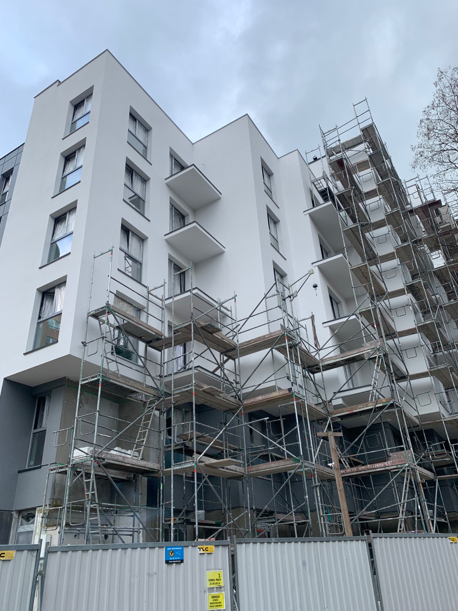 Nowoczesny, biały budynek wielorodzinny w trakcie prac elewacyjnych, otoczony rusztowaniami. Widoczne balkony i okna z zasłonami. Ogrodzenie budowy w dolnej części kadru.