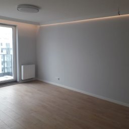 Rem-Instal Michał Laskowski - Pusty pokój po remoncie z drewnianą podłogą, szarymi ścianami, grzejnikiem pod oknem balkonowym i ledowym oświetleniem podsufitowym.