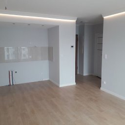 Rem-Instal Michał Laskowski - Pomalowane na szaro wnętrze mieszkania z drewnianą podłogą, widoczne instalacje elektryczne i wodne w ścianie, oraz podświetlany sufit.