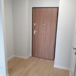 Rem-Instal Michał Laskowski - Wnętrze przedpokoju po remoncie: świeżo pomalowane szare ściany, nowa drewnopodobna podłoga, brązowe drzwi wejściowe z czarną ramą, widoczny fragment białych drzwi wewnętrznych.