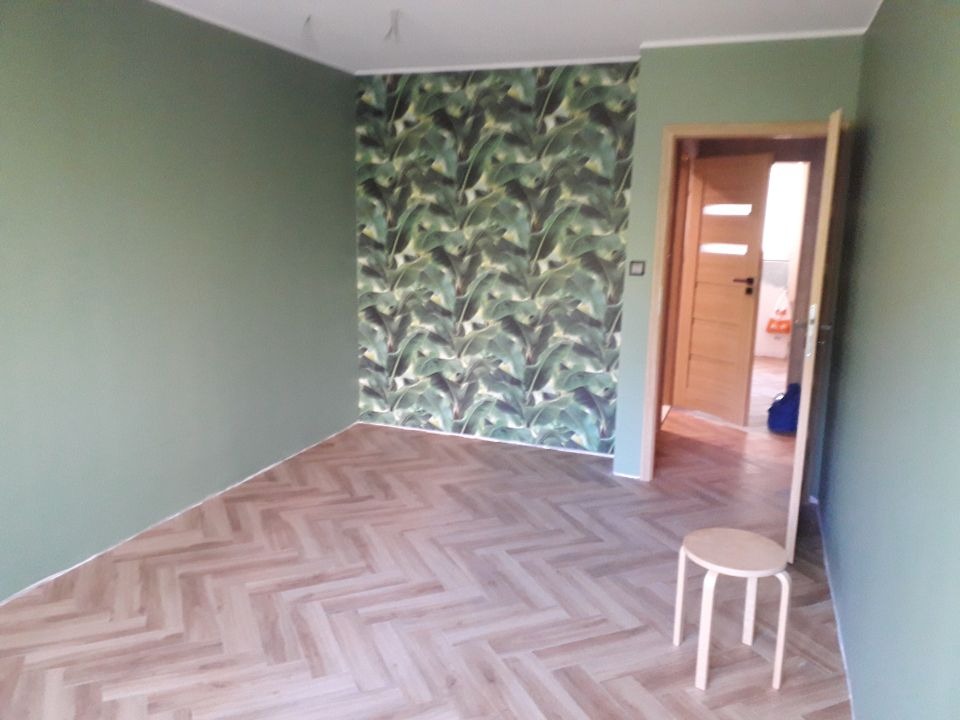 Pomalowane na oliwkowo ściany pomieszczenia z wzorzystą tapetą w motywy liści, parkiet ułożony w jodełkę, widoczny fragment otwartych drzwi i taboret.