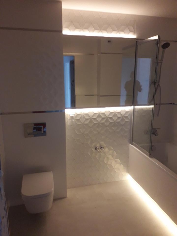 Nowoczesna łazienka z podświetleniem LED pod sufitem i wanną, biała armatura i geometryczna tekstura płytek na ścianie.