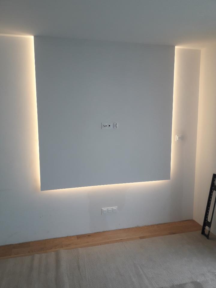 Biała ściana z podświetleniem LED tworzącym ramę, widoczne gniazdka elektryczne, fragment paneli podłogowych i zabezpieczona podłoga.