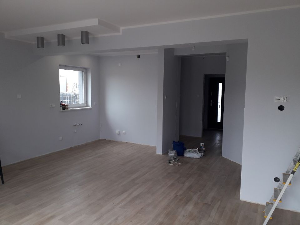 Wnętrze pomalowanego na szaro pokoju z drewnianą podłogą, widoczne puszki elektryczne na ścianie, drabina oparta o ścianę, torba i narzędzia malarskie na podłodze.