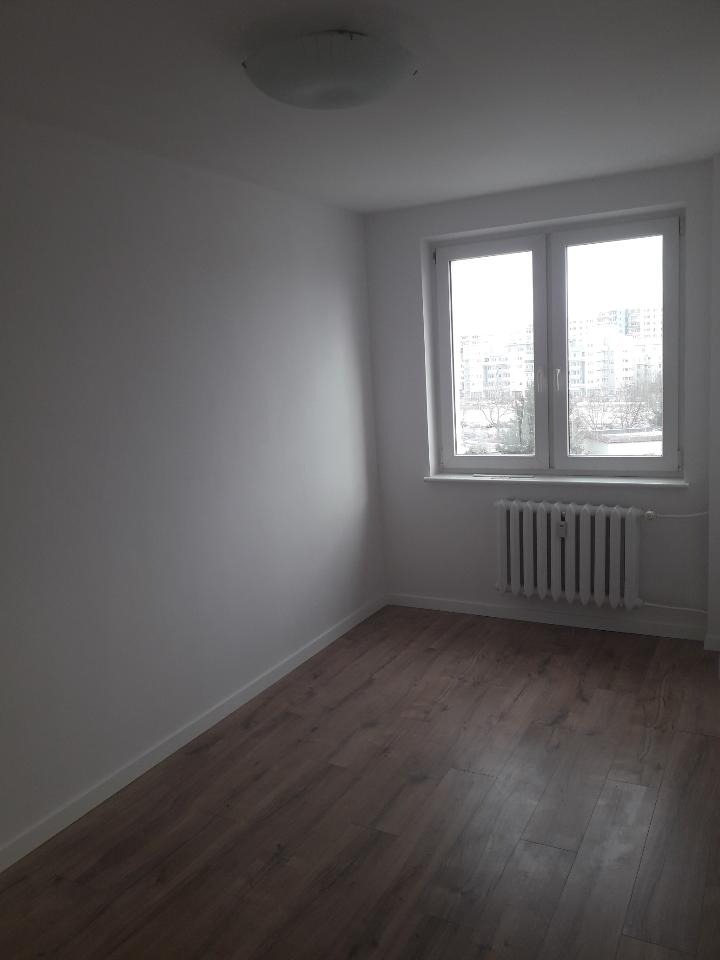 Pusty pokój po remoncie z panelami podłogowymi, świeżo pomalowanymi ścianami na biało, grzejnikiem pod oknem i lampą sufitową.