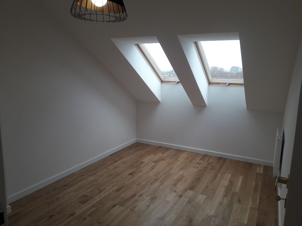 Poddasze po remoncie: świeżo pomalowane białe ściany, drewniana podłoga, dwa okna dachowe w skośnym dachu, nowoczesna lampa wisząca z metalową konstrukcją.