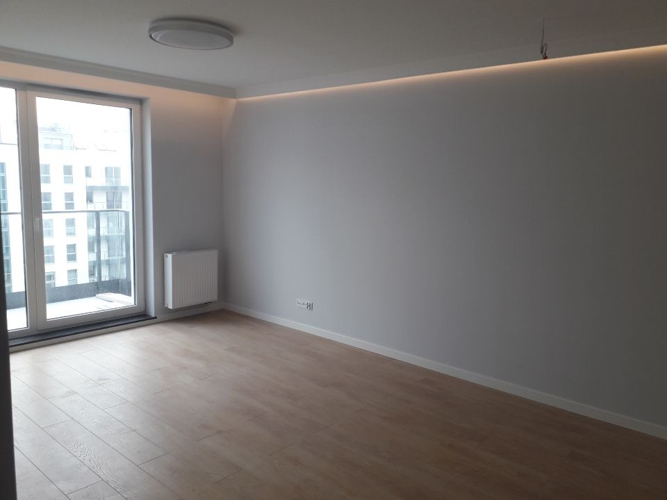 Pusty pokój po remoncie z drewnianą podłogą, szarymi ścianami, grzejnikiem pod oknem balkonowym i ledowym oświetleniem podsufitowym.