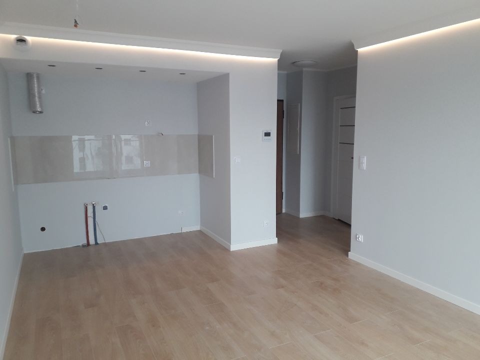 Pomalowane na szaro wnętrze mieszkania z drewnianą podłogą, widoczne instalacje elektryczne i wodne w ścianie, oraz podświetlany sufit.