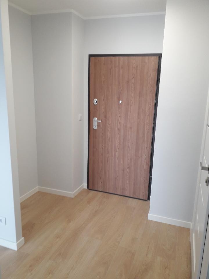 Wnętrze przedpokoju po remoncie: świeżo pomalowane szare ściany, nowa drewnopodobna podłoga, brązowe drzwi wejściowe z czarną ramą, widoczny fragment białych drzwi wewnętrznych.
