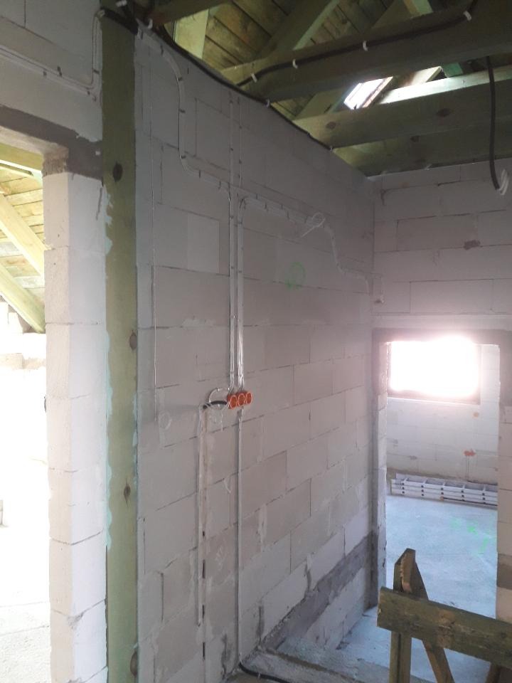 Surowe wnętrze w trakcie prac wykończeniowych: widoczne białe ściany z bloczków, drewniana konstrukcja dachu, poprowadzone kable elektryczne w peszlach i pomarańczowa puszka elektryczna, całość...