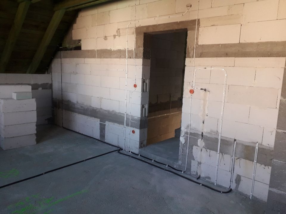 Surowy stan poddasza z widoczną instalacją elektryczną natynkową, przewody w peszlach prowadzone wzdłuż ścian z bloczków betonowych, widoczne puszki instalacyjne, fragment drewnianej konstrukcji...