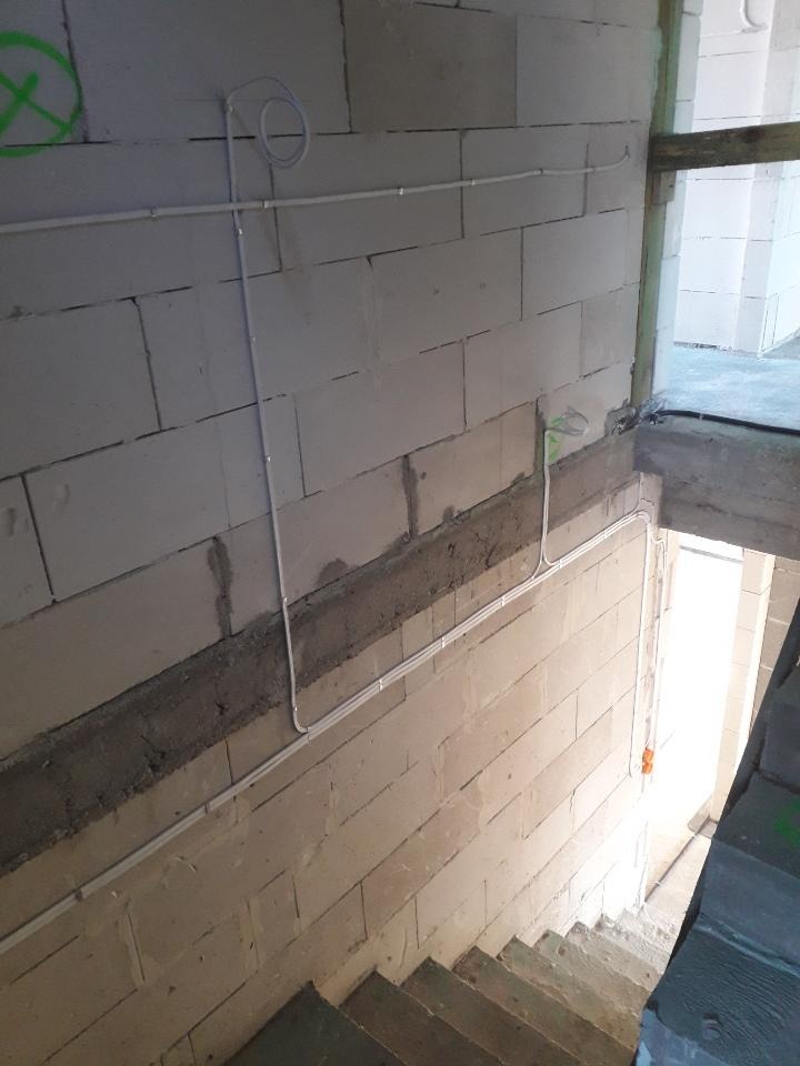 Kable elektryczne prowadzone po ścianie z bloczków betonowych na klatce schodowej w trakcie budowy, widoczne mocowania przewodów i zielony znak na ścianie.
