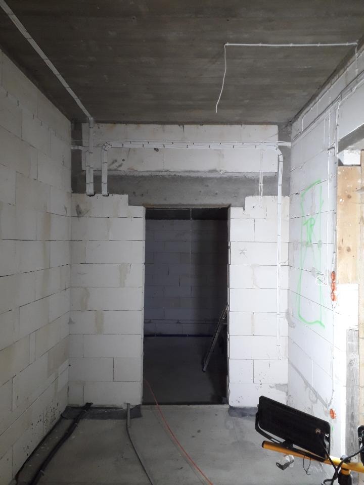 Surowy stan wnętrza z białych bloczków, widoczne kable elektryczne prowadzone po ścianach i suficie, tymczasowe oświetlenie budowlane na statywie, zielone oznaczenia na ścianie.