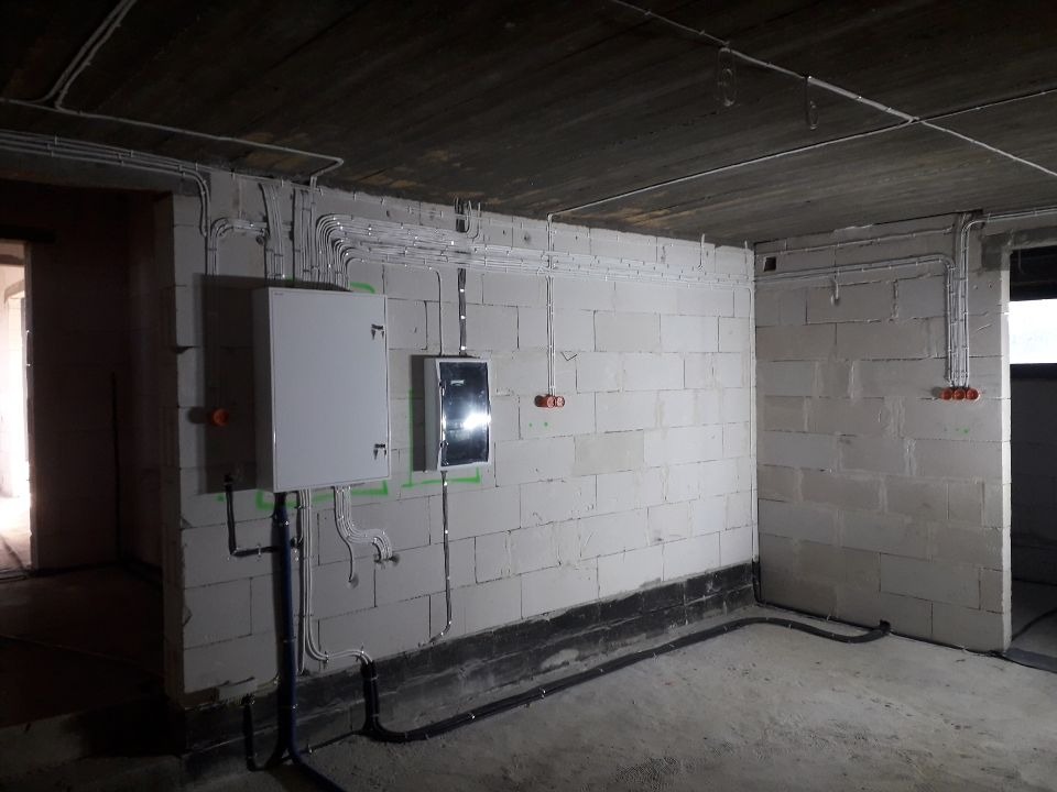 Surowy stan instalacji elektrycznej w pomieszczeniu z betonowym sufitem i ścianami z białego bloczka, widoczne kable w peszlach, puszki instalacyjne i dwie szafy rozdzielcze.