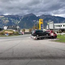 Czarny samochód osobowy na czerwonej lawecie na parkingu stacji benzynowej JET, w tle góry i pochmurne niebo.