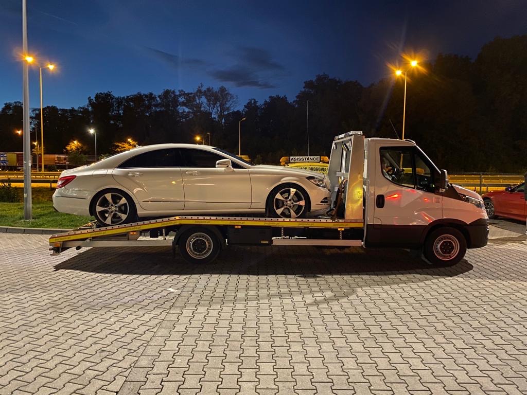 Biały samochód osobowy marki Mercedes-Benz CLS na platformie holowniczej lawety, na parkingu wyłożonym kostką, widoczne światła uliczne i drzewa w tle, nocne ujęcie.