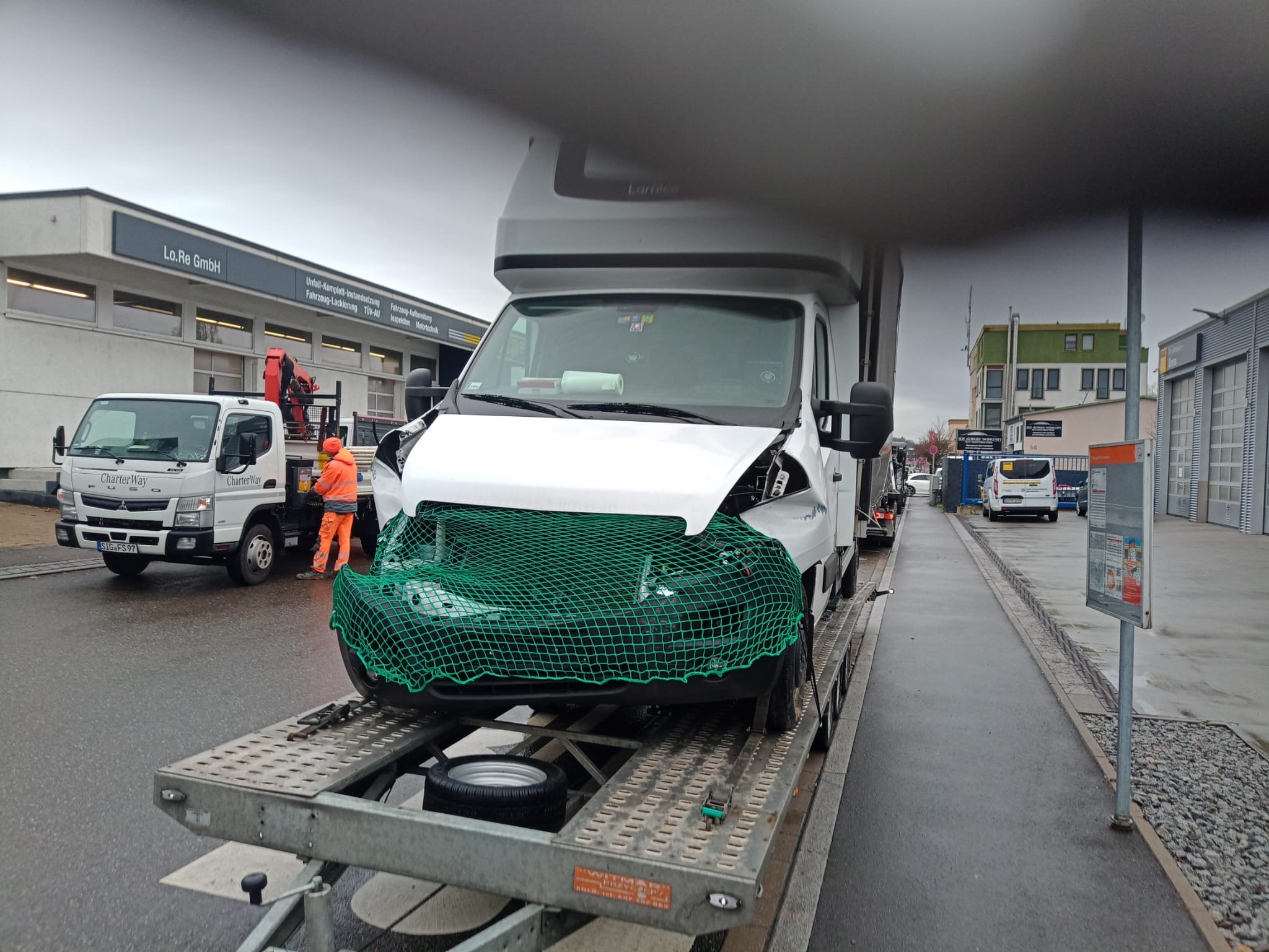 Uszkodzony biały samochód dostawczy zabezpieczony zieloną siatką, umieszczony na lawecie. W tle budynek warsztatu samochodowego i pracownik w pomarańczowym kombinezonie przy mniejszej lawecie.