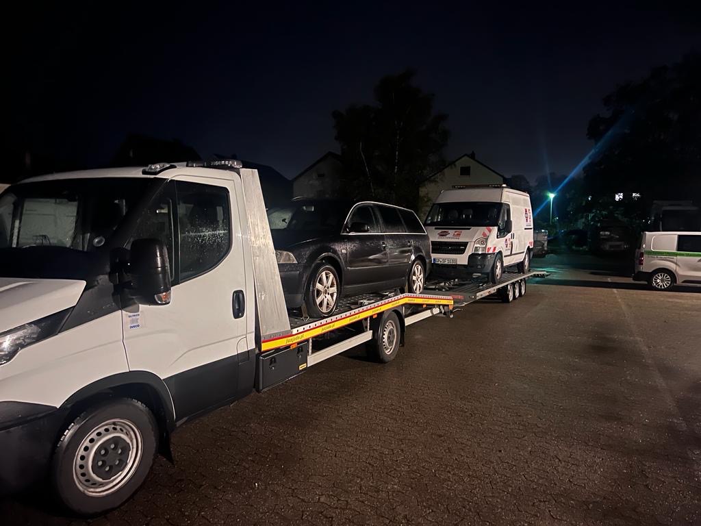 Czarny samochód osobowy kombi na białej lawecie w nocy, obok zaparkowany biały bus z pomarańczowymi elementami graficznymi.