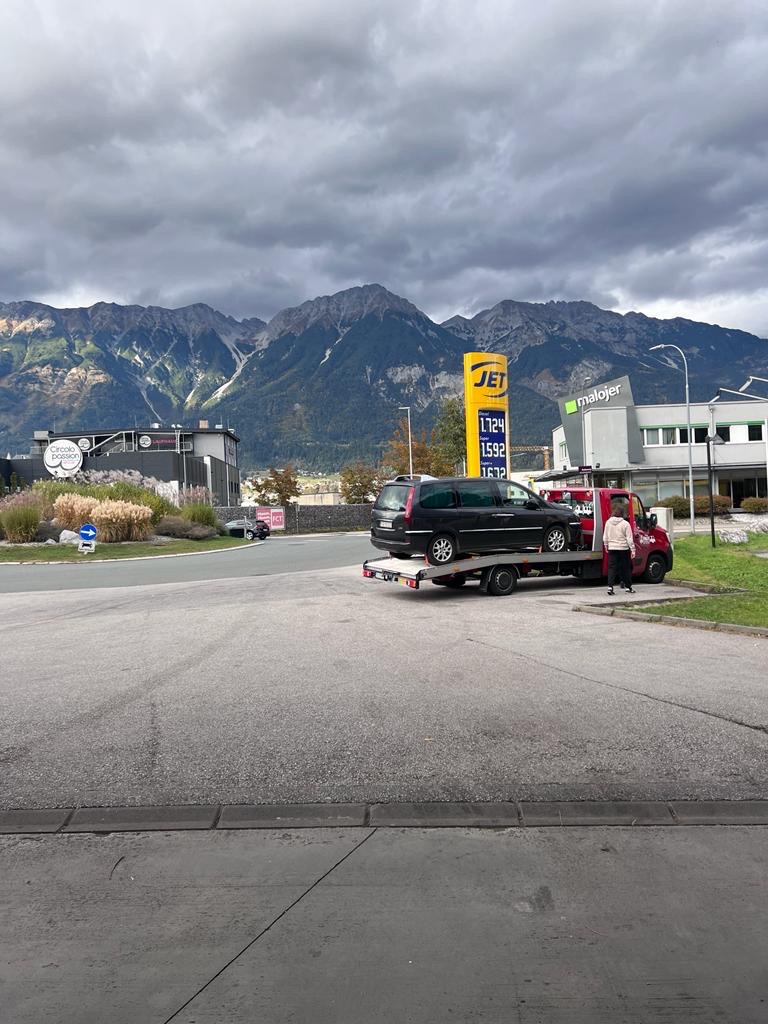 Czarny samochód osobowy na czerwonej lawecie na parkingu stacji benzynowej JET, w tle góry i pochmurne niebo.