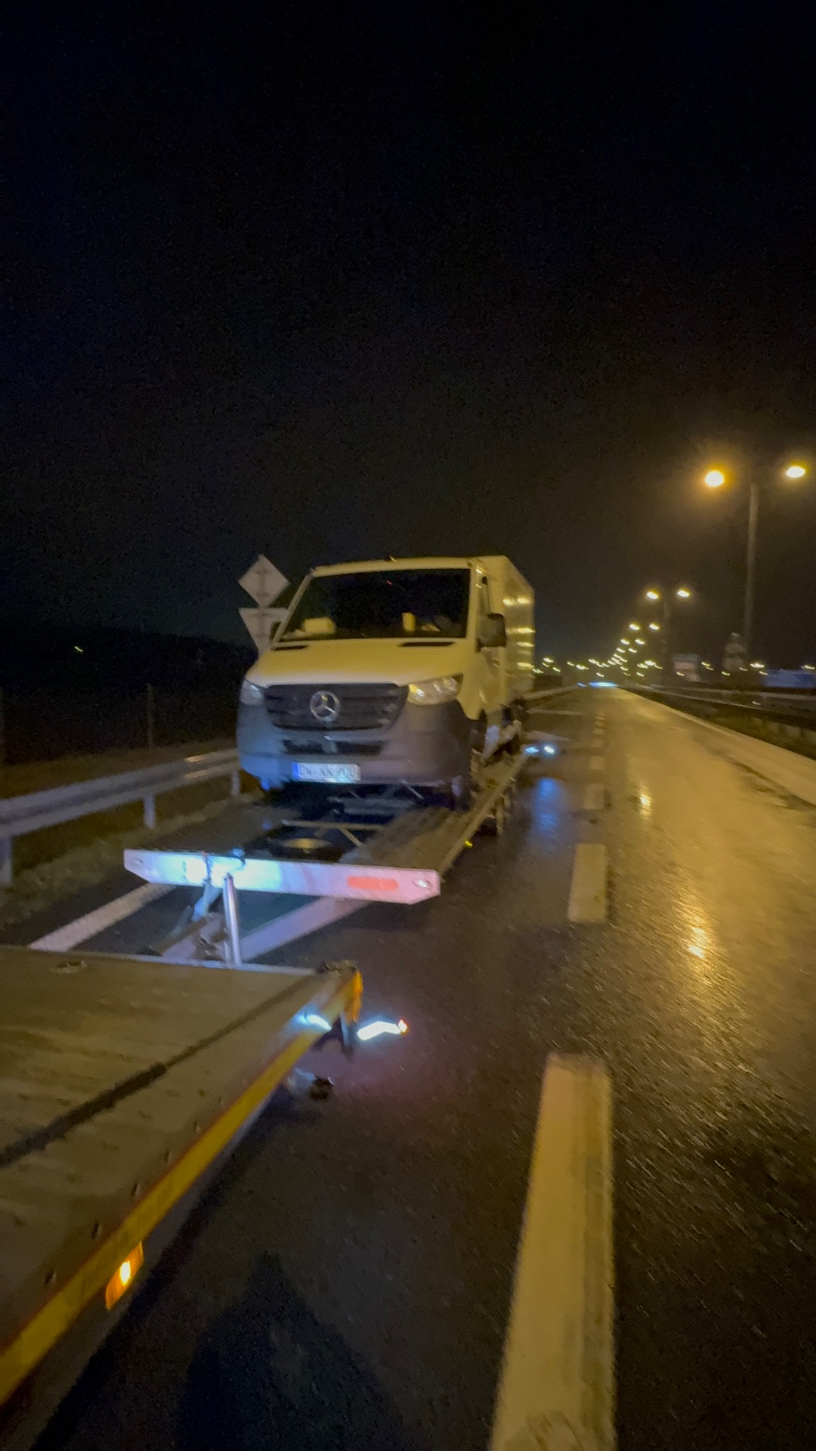 Biały samochód dostawczy marki Mercedes przewożony na platformie lawety na mokrej, oświetlonej ulicy nocą.