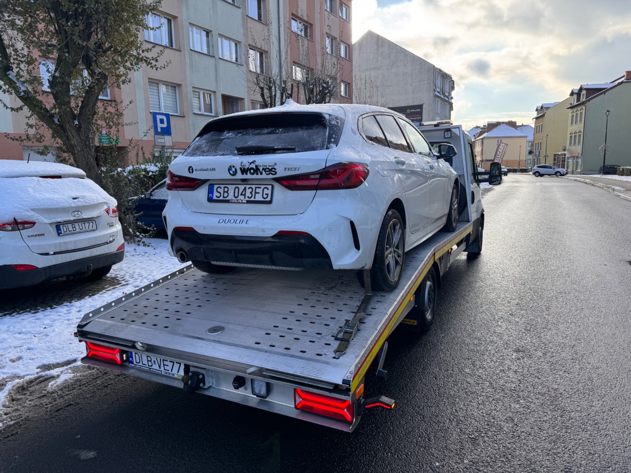 Białe BMW 118i na lawecie w zimowej scenerii miejskiej, widok z tyłu, tablice rejestracyjne SB 043FG i DLB-VE77, naklejki 'wolves' i 'DUOLIFE'.