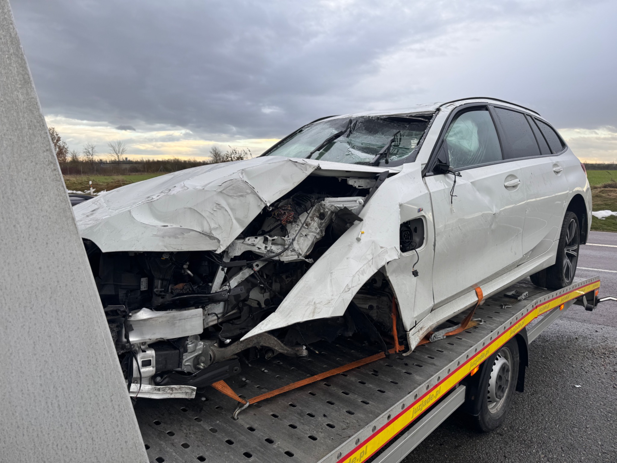 Uszkodzony, biały samochód osobowy BMW na platformie lawety, zabezpieczony pomarańczowymi pasami. Zniszczony przód pojazdu, pęknięta szyba czołowa. Pochmurne niebo w tle.
