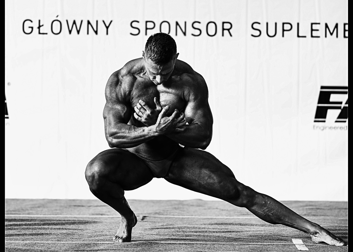 Czarno-białe zdjęcie muskularnego mężczyzny w pozycji przysiadu na scenie, z widocznym logo sponsora w tle. Wyraźna definicja mięśni i ostre światło.