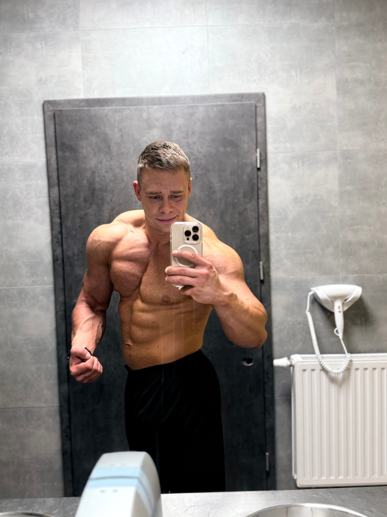 Mężczyzna z imponującą muskulaturą pozuje do selfie w szarej łazience, prezentując wyrzeźbione ciało i trzymając telefon w dłoni.