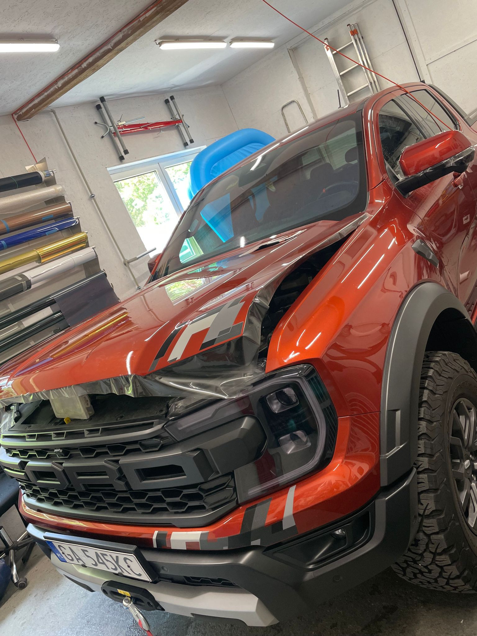 Pomarańczowy Ford Ranger Raptor w trakcie oklejania folią ochronną, widoczne rolki folii w tle.