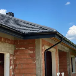 Nowy dom w trakcie budowy z czarną blachą trapezową na dachu i systemem rynnowym, widoczne okna bez szyb i drabina oparta o ścianę.