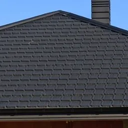 Nowy dach pokryty czarnym gontem bitumicznym, komin z blachy w kolorze antracytowym, widoczny fragment surowej ściany budynku z czerwonej cegły.