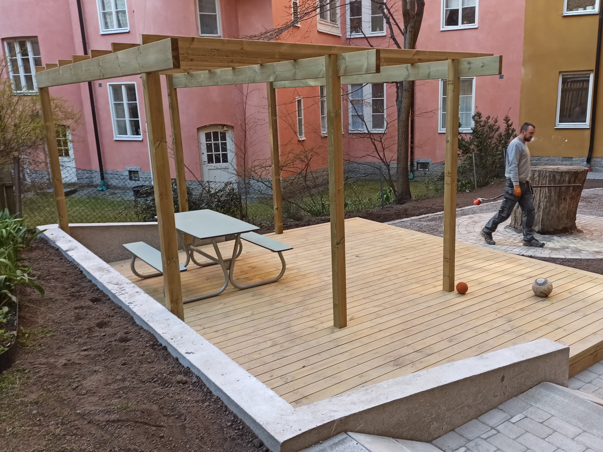 Nowo wybudowany taras z jasnego drewna z drewnianą pergolą, otoczony betonową obwódką i świeżo przygotowanym terenem, z metalowym stołem piknikowym i piłkami na desce, na tle różowych budynków...