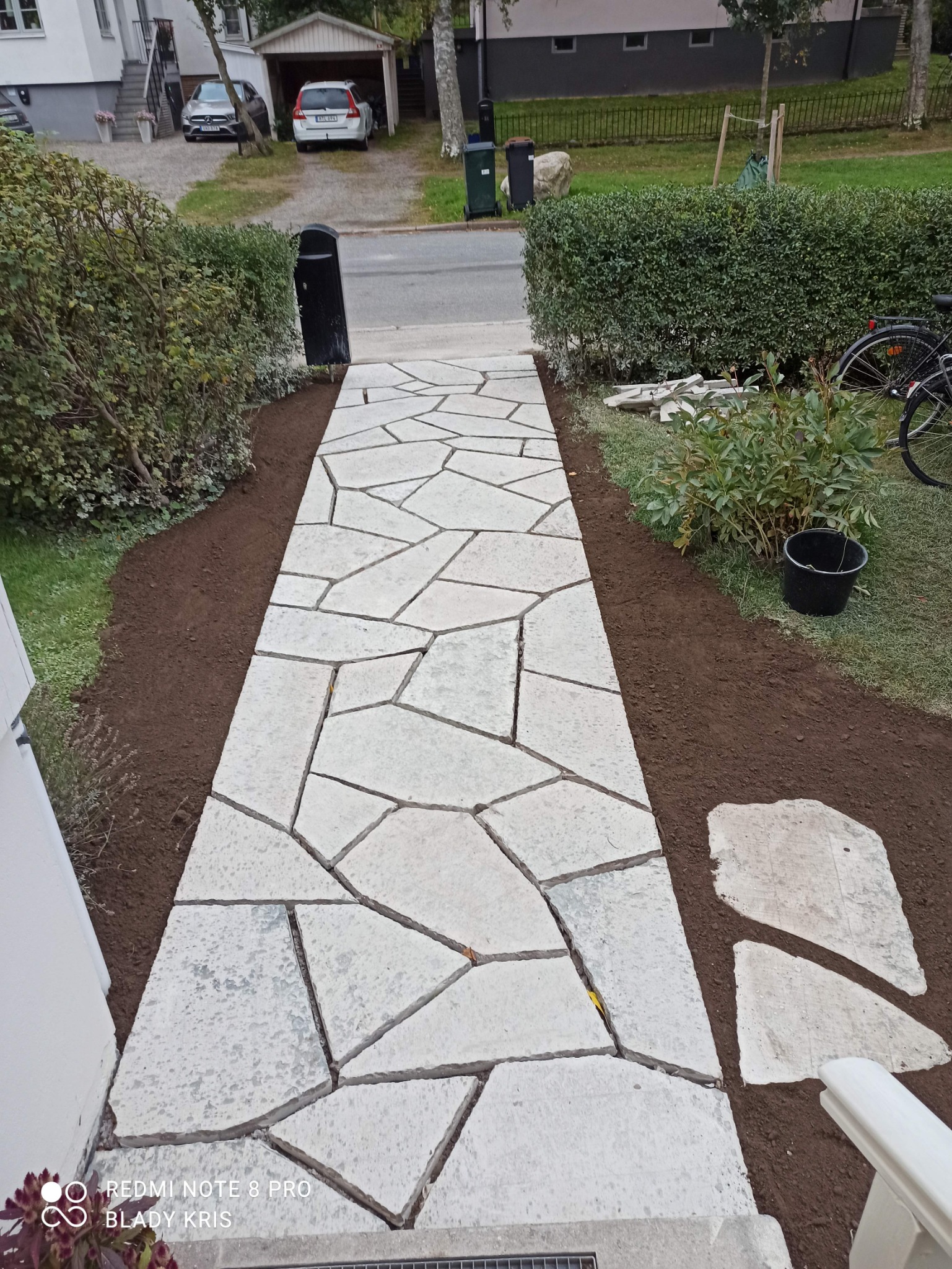 Ścieżka z nieregularnych płyt kamiennych prowadząca do domu, otoczona świeżo przekopaną ziemią i zielenią, widok z góry.