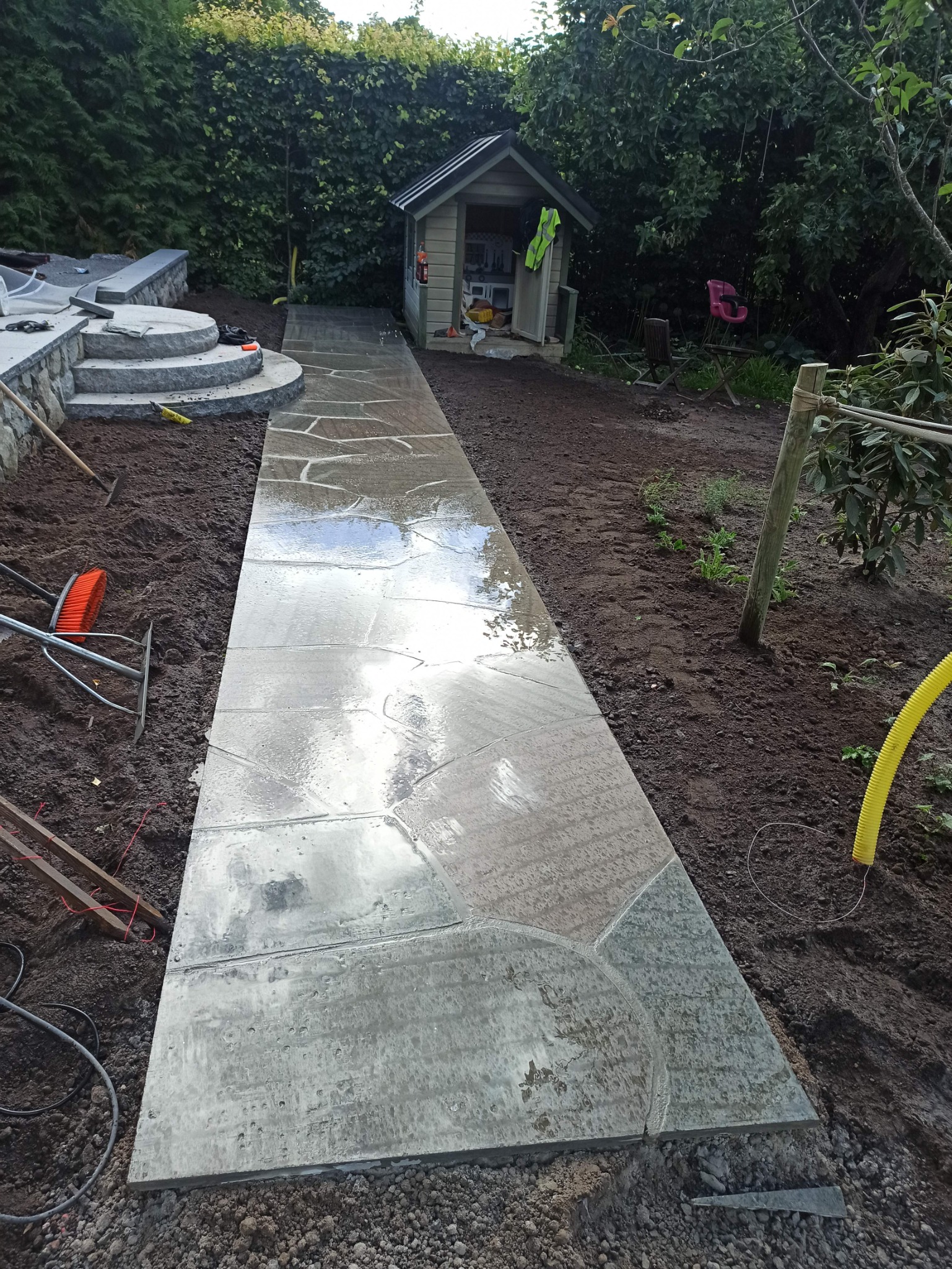 Świeżo ułożony, mokry kamienny chodnik w ogrodzie, prowadzący do małego domku narzędziowego; widoczne narzędzia ogrodnicze i przygotowane podłoże.