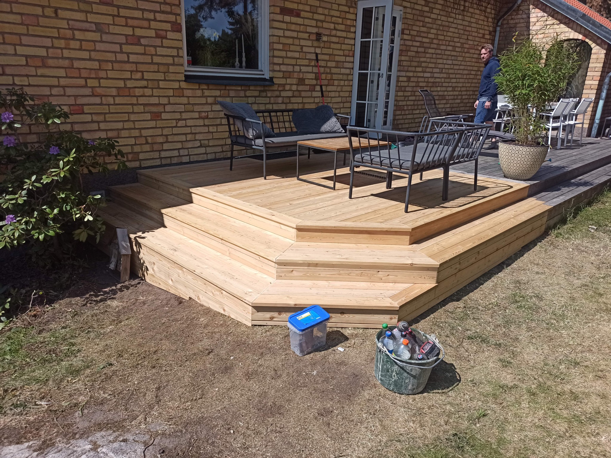 Nowo wybudowany, jasny, drewniany taras z kilkoma poziomami schodów, wyposażony w szare meble ogrodowe i rośliny w donicach, na tle ceglanego domu. W tle widoczny mężczyzna i dodatkowe krzesła.