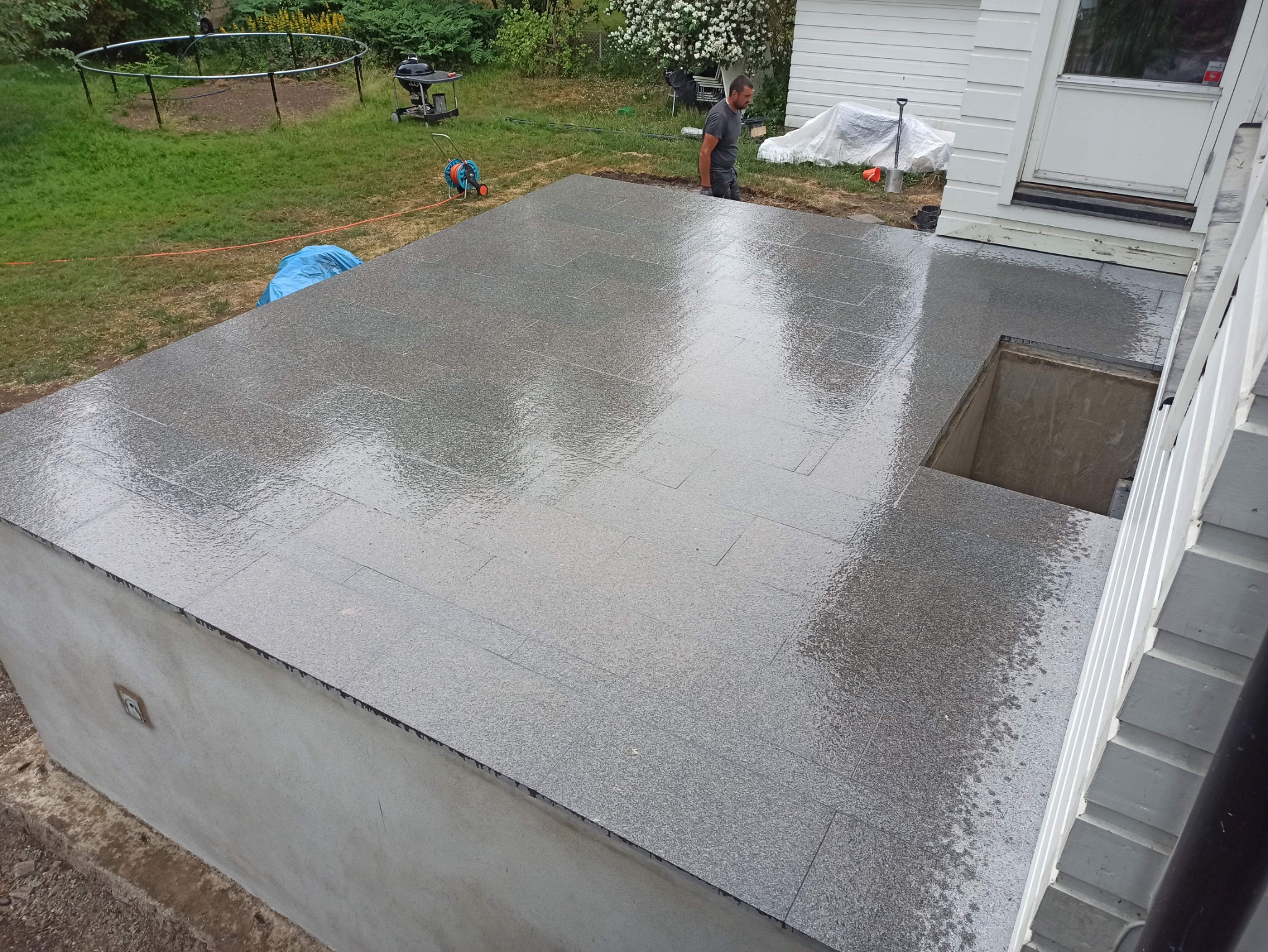 Świeżo ułożony taras z szarych, kamiennych płyt po deszczu, przylegający do białego domu z widocznym włazem do piwnicy i pracującym mężczyzną w tle.
