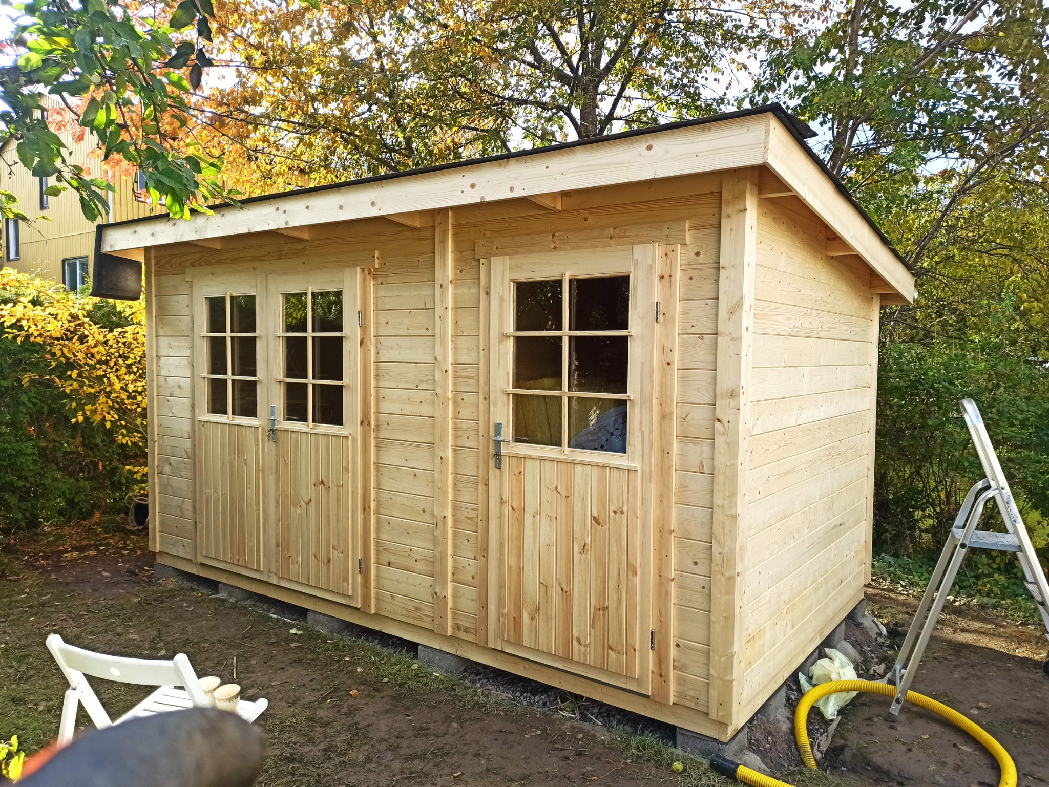 Nowo postawiony, drewniany domek narzędziowy z dwoma oknami i drzwiami, widoczna drabina oparta o ścianę, żółty wąż i krzesło ogrodowe w tle drzew i krzewów.
