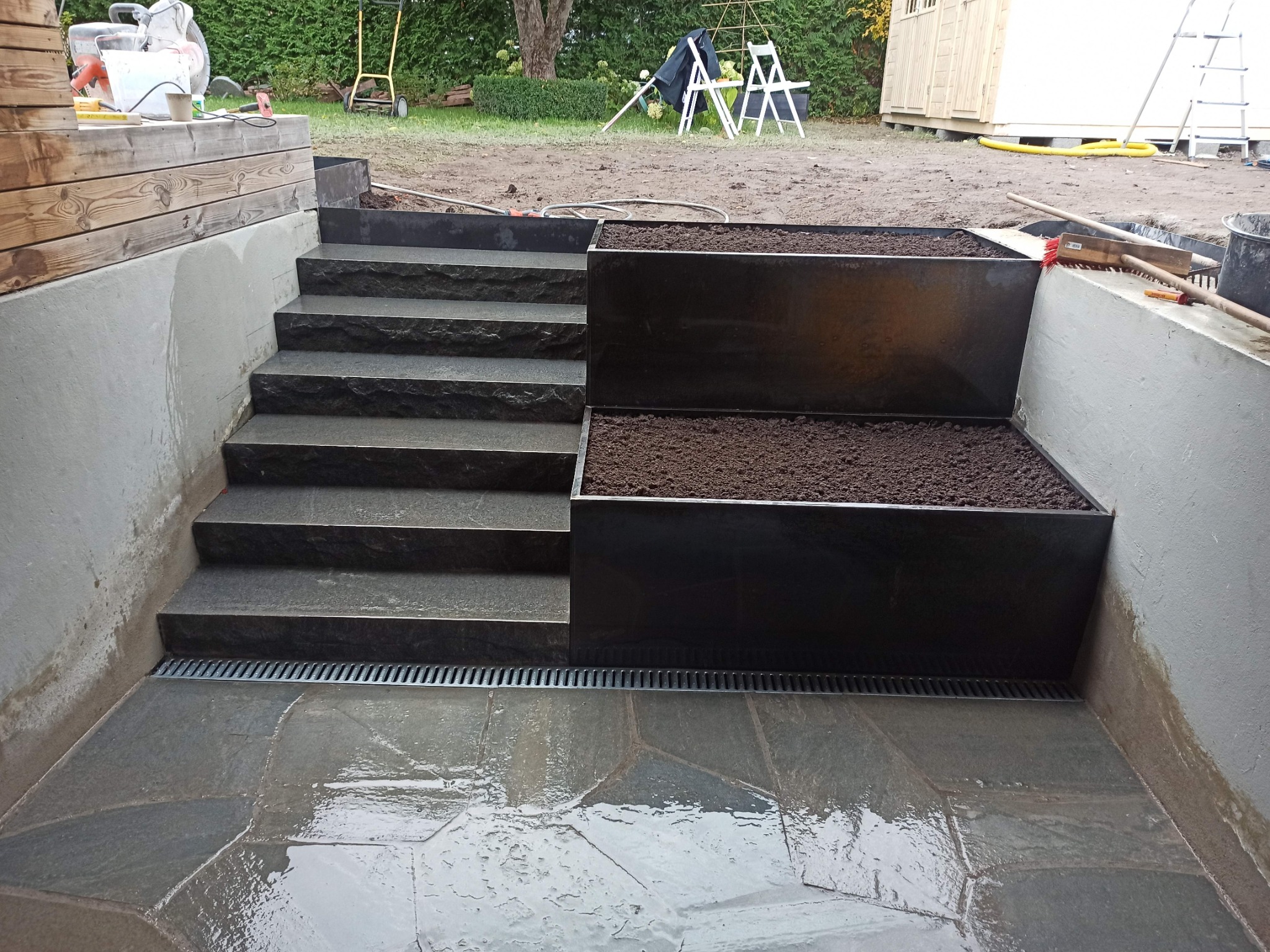 Wykończony taras z kamiennymi płytami w nieregularnym kształcie, schody z ciemnego kamienia prowadzące w górę, nowoczesne metalowe donice wypełnione ziemią, widoczny system odprowadzania wody.