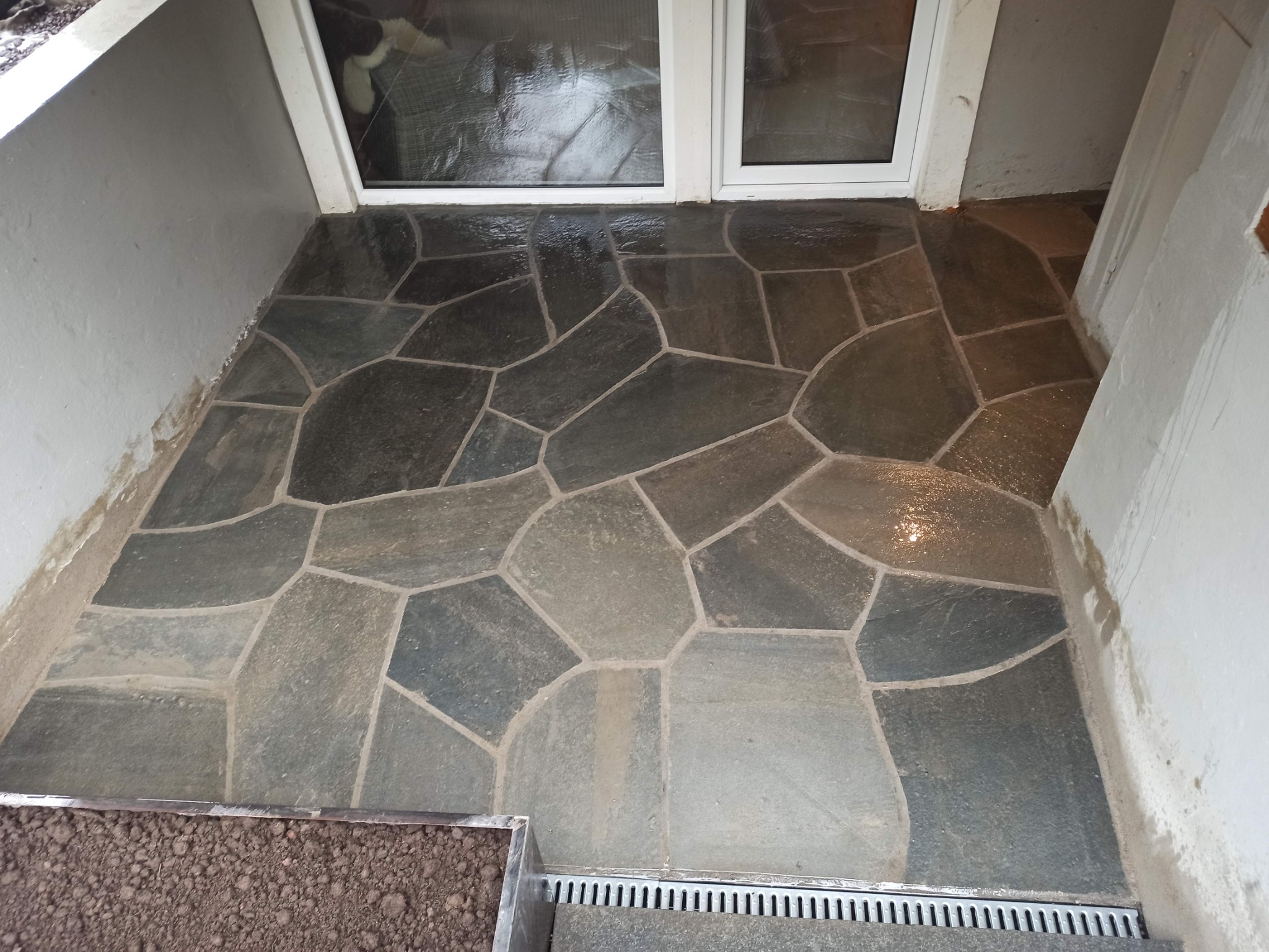 Nieregularne płyty kamienne tworzące taras przed wejściem do budynku, widoczne ślady zaprawy, obok kratka odpływowa i pojemnik z ziemią.