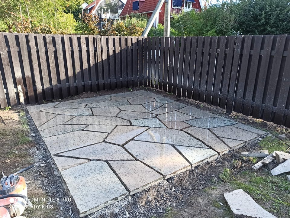 Nowo ułożony taras z nieregularnych płyt kamiennych, przylegający do ciemnego, drewnianego ogrodzenia. W tle widoczny dom z czerwonym dachem.