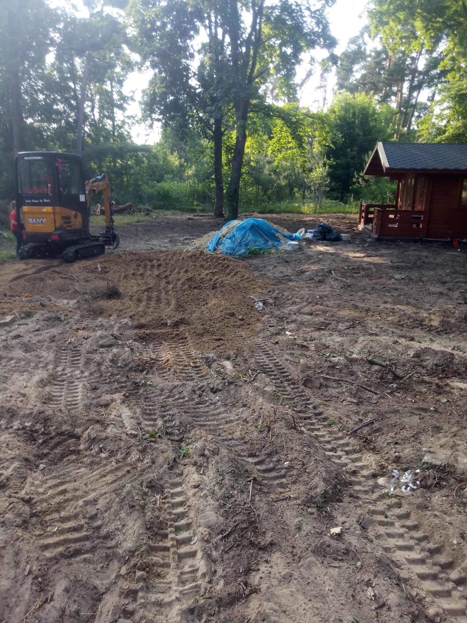 Teren przygotowany pod budowę, z widocznym mini-koparką SANY, wyrównaną ziemią z wyraźnymi śladami gąsienic, stertą ziemi przykrytą niebieską plandeką oraz drewnianą altaną w tle.