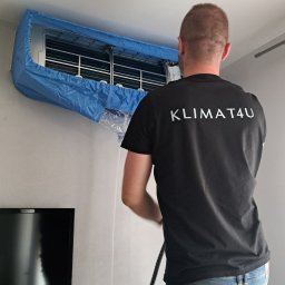 KLIMAT4U - Mężczyzna na drabinie czyści klimatyzator domowy, zabezpieczony niebieską osłoną, z logo firmy 'KLIMAT4U' na czarnej koszulce.