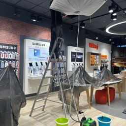 KLIMAT4U - Pracownik na drabinie montuje lub naprawia instalację podwieszaną pod sufitem w salonie sprzedaży, zabezpieczone folią meble, zielony i niebieski pojemnik oraz urządzenie do czyszczenia.