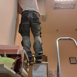 KLIMAT4U - Pracownik w firmowej koszulce KLIMATA4U stoi na szafce, naprawiając lub instalując coś w suficie podwieszanym, widoczne narzędzia i drabina aluminiowa w tle.