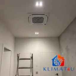 Kasetonowa jednostka klimatyzacji zamontowana w suficie podwieszanym, widoczna z dołu, z drabinką rozstawną opartą o ścianę i logo firmy w prawym dolnym rogu.