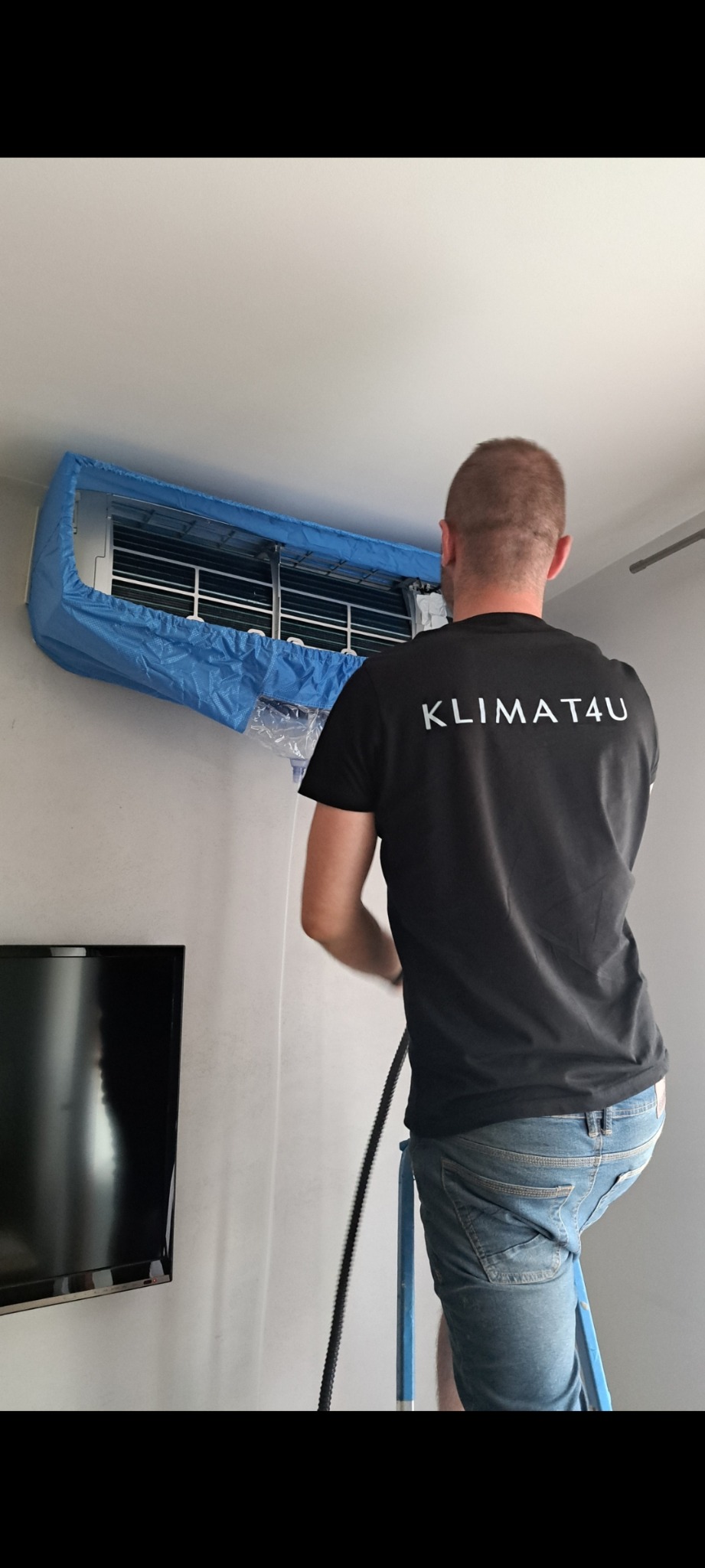 Mężczyzna na drabinie czyści klimatyzator domowy, zabezpieczony niebieską osłoną, z logo firmy 'KLIMAT4U' na czarnej koszulce.